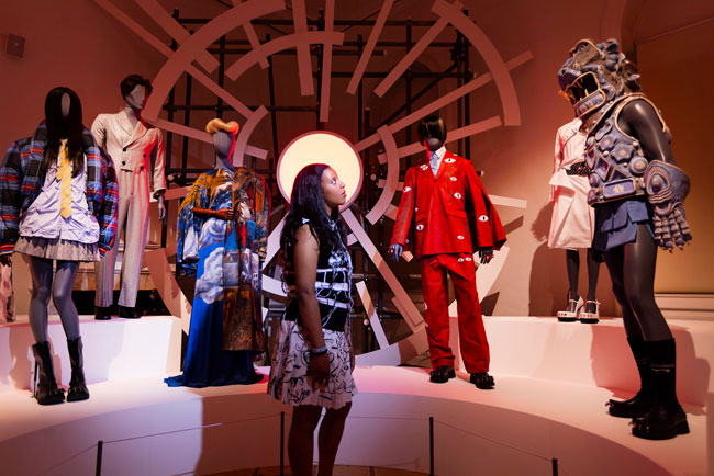 A young woman in a gallery space looking at a raised plinth with mannequins wearing iconic LOVERBOY outfits. The backdrop is a large, lit circular sphere with lighting elements radiating outwards