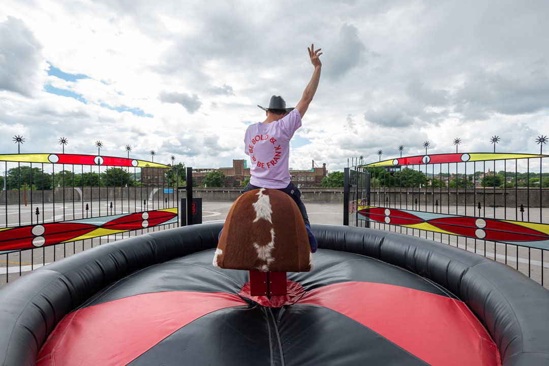 A man with his back to the camera rides a mechanical bull. He wears a cowboy hat and a light-colored t-shirt with indiscernible text. His left arm is raised in the air, and he appears to be trying to maintain his balance.