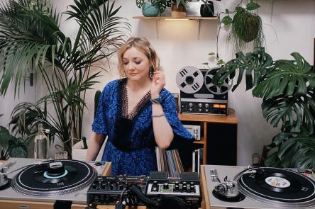 Alena Arples is at work on the decks. She is DJing in a light-filled room full of plants.