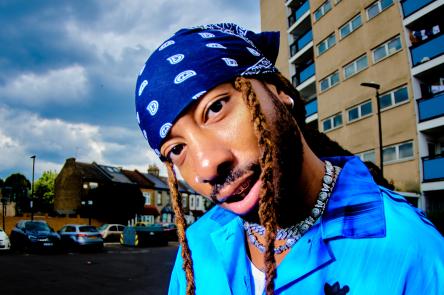 Donnie Sunshine wears a blue headscarf with two braids framing his face. They are wearing a blue adidas shirt.