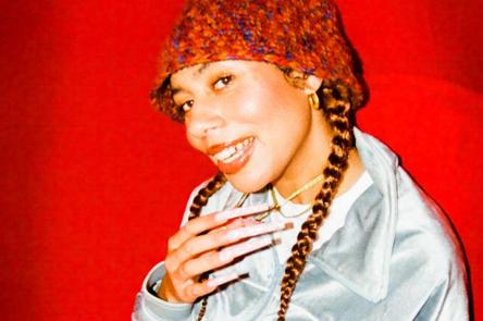 A portrait of DJ and singer Ella Knight in a textured bucket hat, sitting in a red chair