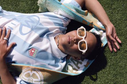 GIN lays on the grass with the camera shooting them from above. They are bathed in sunshine and smiling, while wearing a football shirt and white sunglasses.