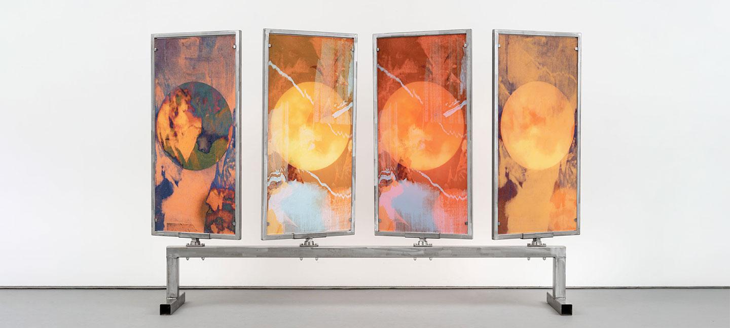 Four rectangular panels, framed in silver metal, are mounted on a long horizontal bar supported by two legs. Each panel displays an abstract image dominated by a central circular shape reminiscent of a celestial body.