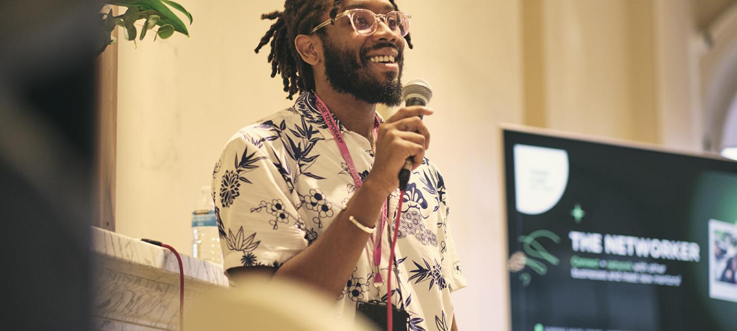 A photo of Akil Benjamin leading a workshop as part of the Black Business Incubator. Akil is Black man with medium length dreads and a beard. He is smiling and holding a microphone.