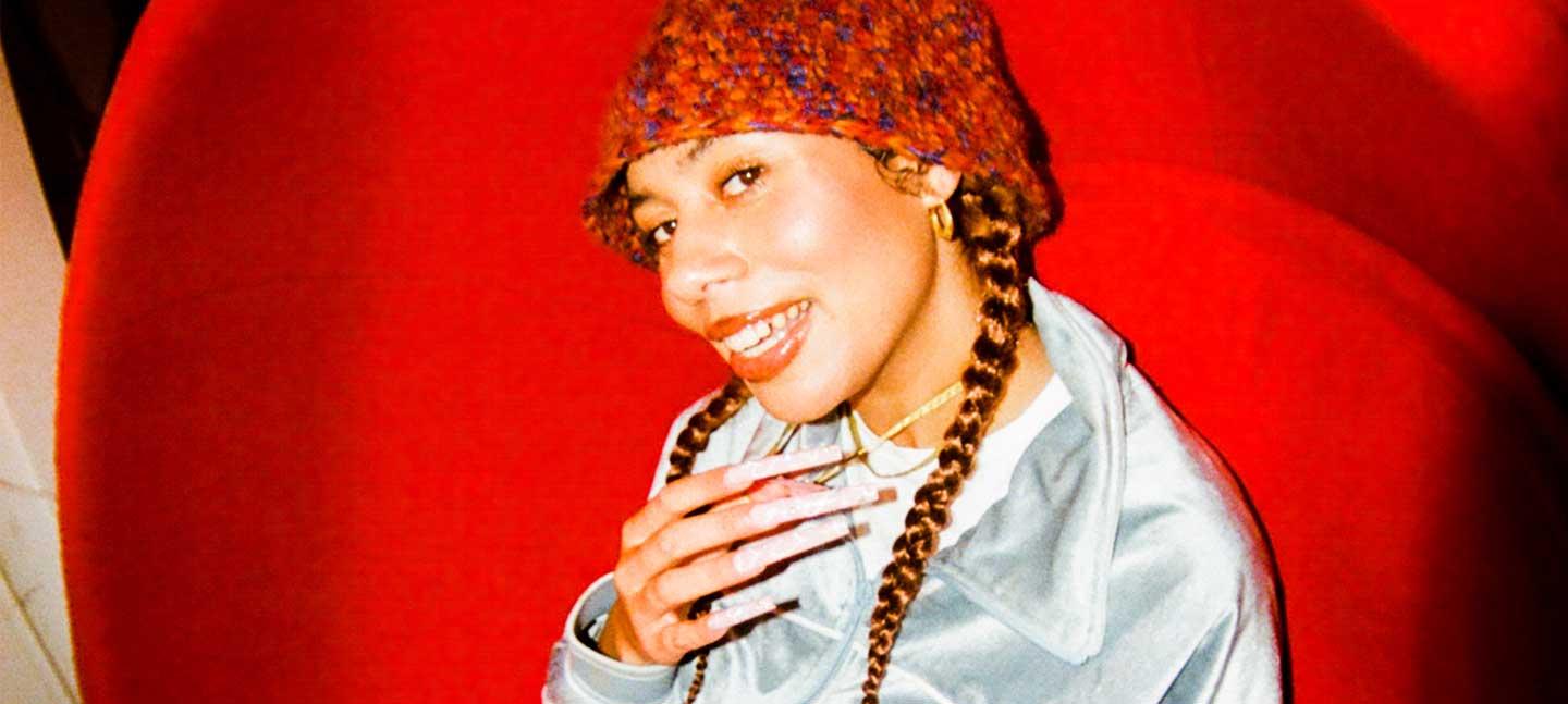 A portrait of DJ and singer Ella Knight in a textured bucket hat, sitting in a red chair