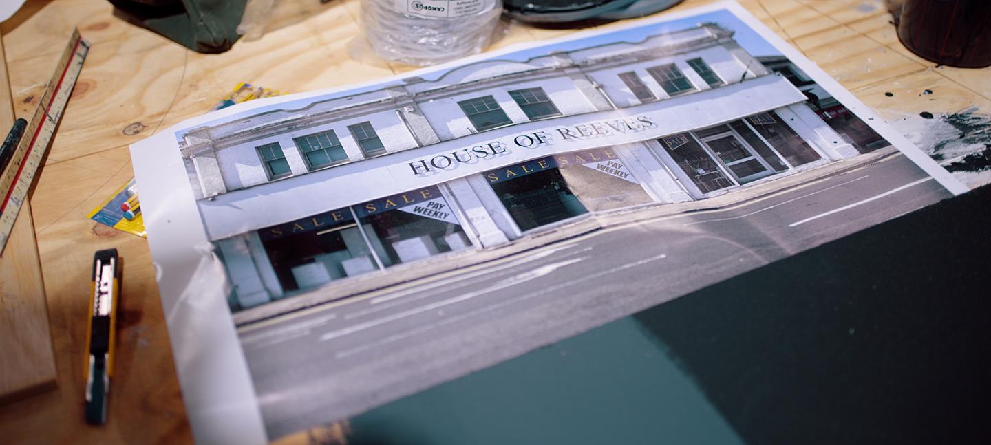 A photo of the furniture shop House of Reeves lays on a desk, surrounded by paint stains, a stanley knife and bottle of white spirits.