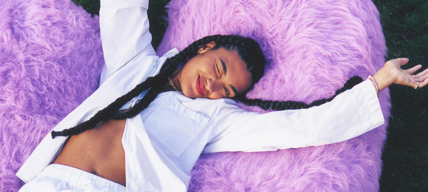 Musician Olivia Dean winks cheekliy at the camera as she lies on a giant violet fur heart, dressed in a white linen suit