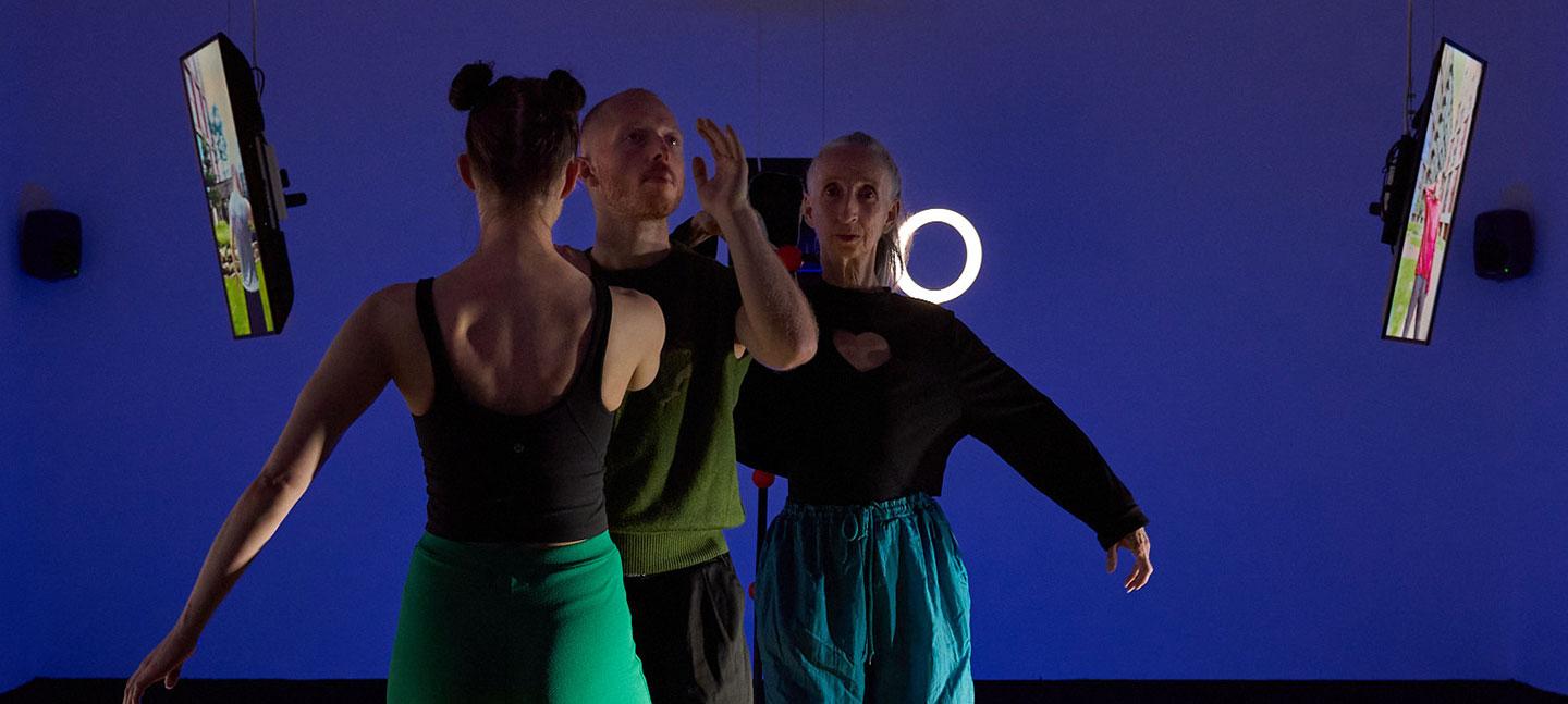 Three people stand facing away from the camera against a vibrant blue wall. On the left, a woman with her hair in two buns wears a black tank top and a green skirt. In the center, a man with short hair wears a green t-shirt.