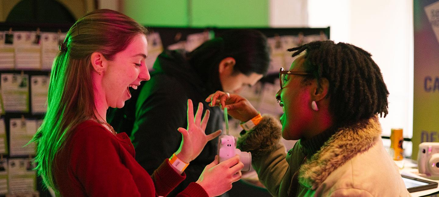 Two women laughing and engaging with a small interactive device at an indoor event