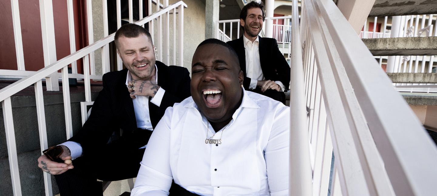 A close portrait shot of Gabriels, with the 3 members seated on a building's fire escape stairs and laughing out loud