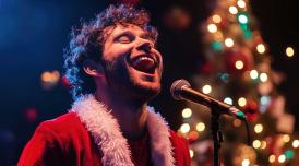 A male performer dressed in a Santa Claus costume sings full voice with his eyes closed. A Christmas tree is in the background