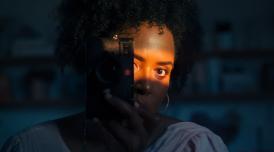Holly-Marie, a Black woman takes a photo of herself in a mirror. The room is in shadow and there is a patch of sunlight lighting up her face. Her face is half obscured by the camera she holds to her eye.