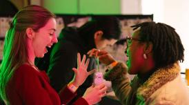 Two women laughing and engaging with a small interactive device at an indoor event