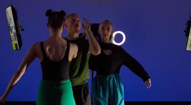 Three people stand facing away from the camera against a vibrant blue wall. On the left, a woman with her hair in two buns wears a black tank top and a green skirt. In the center, a man with short hair wears a green t-shirt.